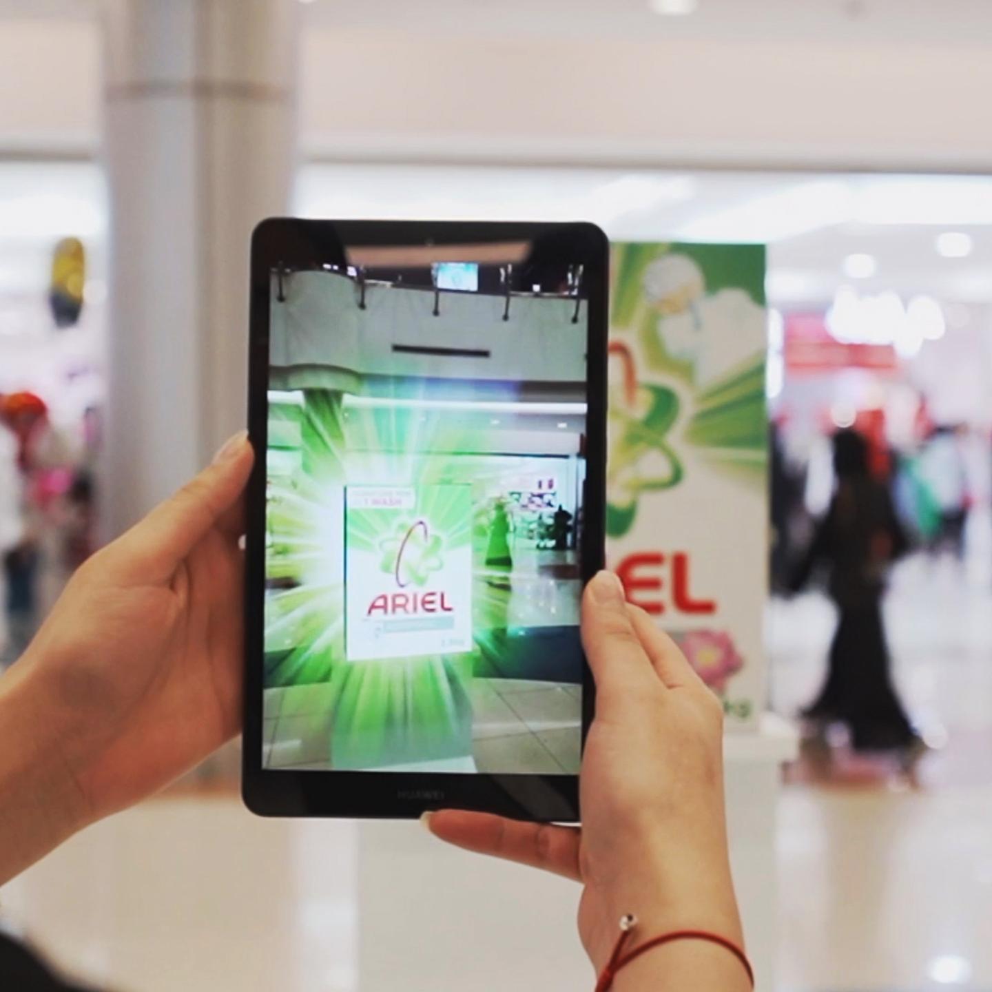 Over-the-shoulder view of a person holding a tablet that displays an augmented reality (AR) animation of the Ariel brand. The tablet camera is directed towards an Ariel advertisement in a shopping mall, where the AR feature overlays engaging graphics and product information onto the real-world image, creating an immersive promotional experience.