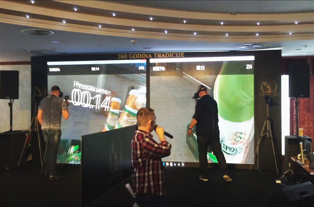 Two competitors in VR headsets sorting virtual beer bottles into corresponding boxes against a timer, with an audience watching their progress on a large screen.