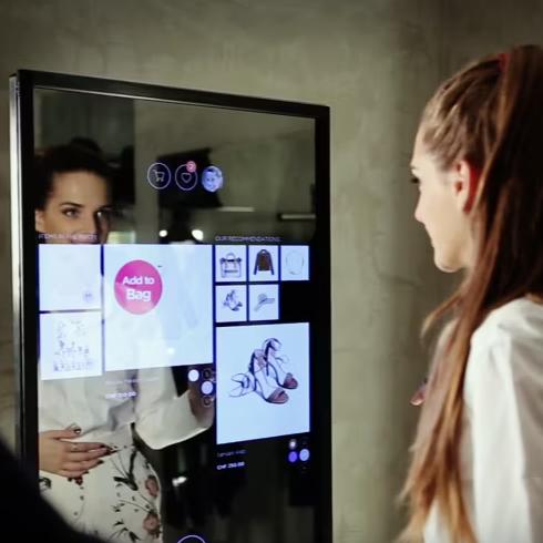 A woman interacts with a high-tech mirror in a fitting room, displaying various clothing items and virtual buttons for a premium shopping experience.