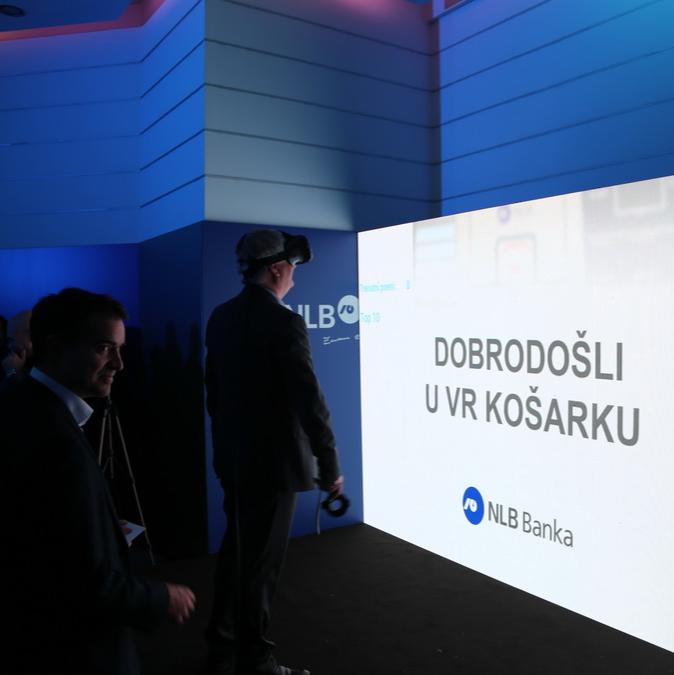 Two men standing in a blue-lit room at a VR event, facing a projection screen with the text "DOBRODOŠLI U VR KOŠARKU" alongside the NLB Banka logo.