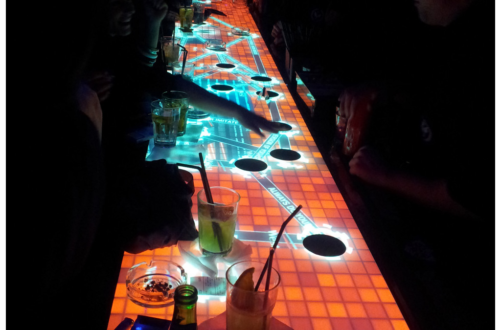Overhead view of an interactive bar surface glowing with neon light patterns, where objects and touch are detected by proprietary infrared sensors.