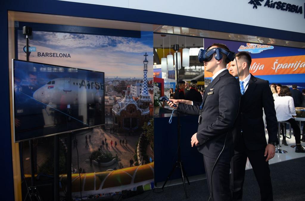 An Air Serbia representative in a pilot's uniform assists a visitor using VR to experience the airline's service, with a backdrop promoting Barcelona, one of their new destinations.