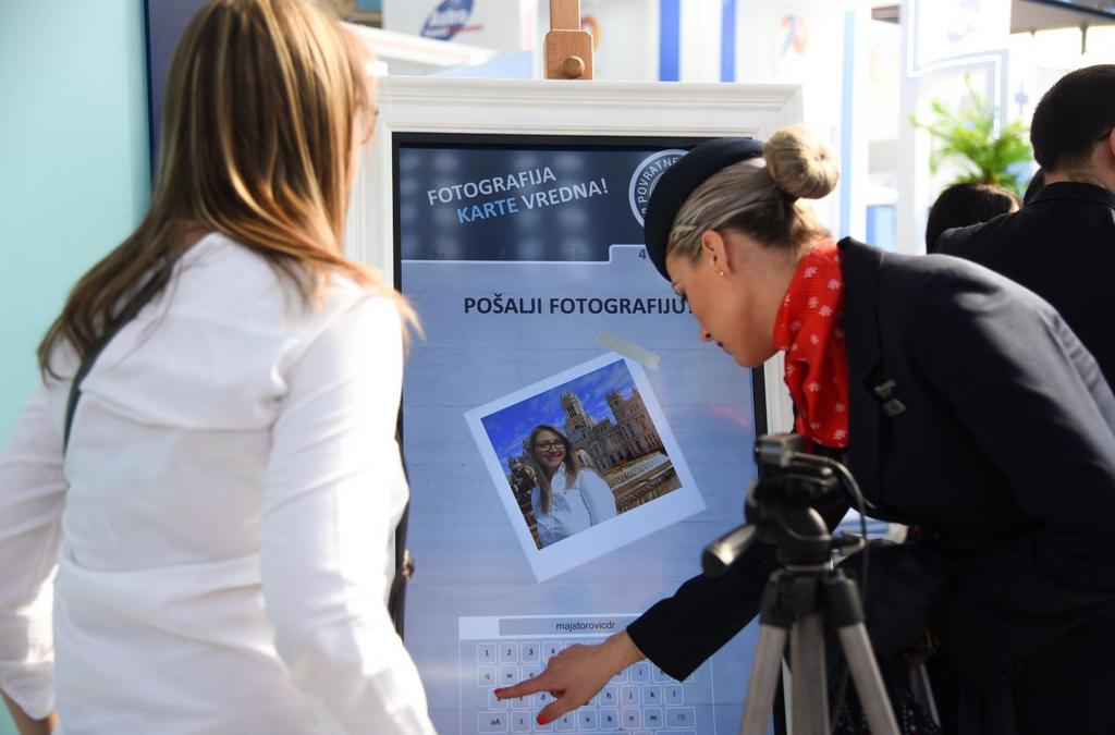 A visitor is engaged with a touchscreen kiosk at Air Serbia's booth, sending a digital postcard, highlighting the airline's new destinations promoted at the Tourism Fair.