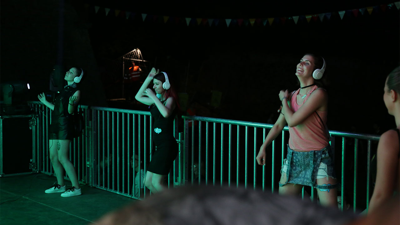 Smiling participants wearing headphones dance in front of the Guarana No Sleep stage, with their silhouettes cast against an illuminated Guarana owl backdrop, showcasing the energetic atmosphere of the EXIT Festival.