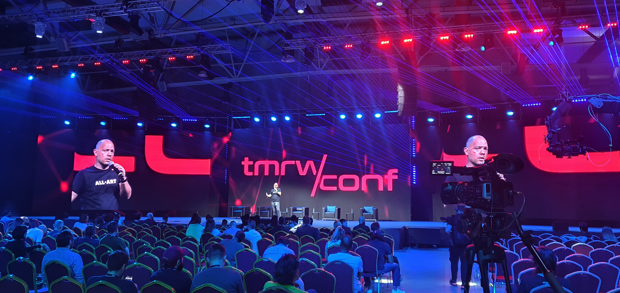 The TMRW Conference session in progress with a speaker on stage discussing the future of digital art, set against a dramatic backdrop of red and blue light beams, highlighting the event's focus on virtual reality and the arts.