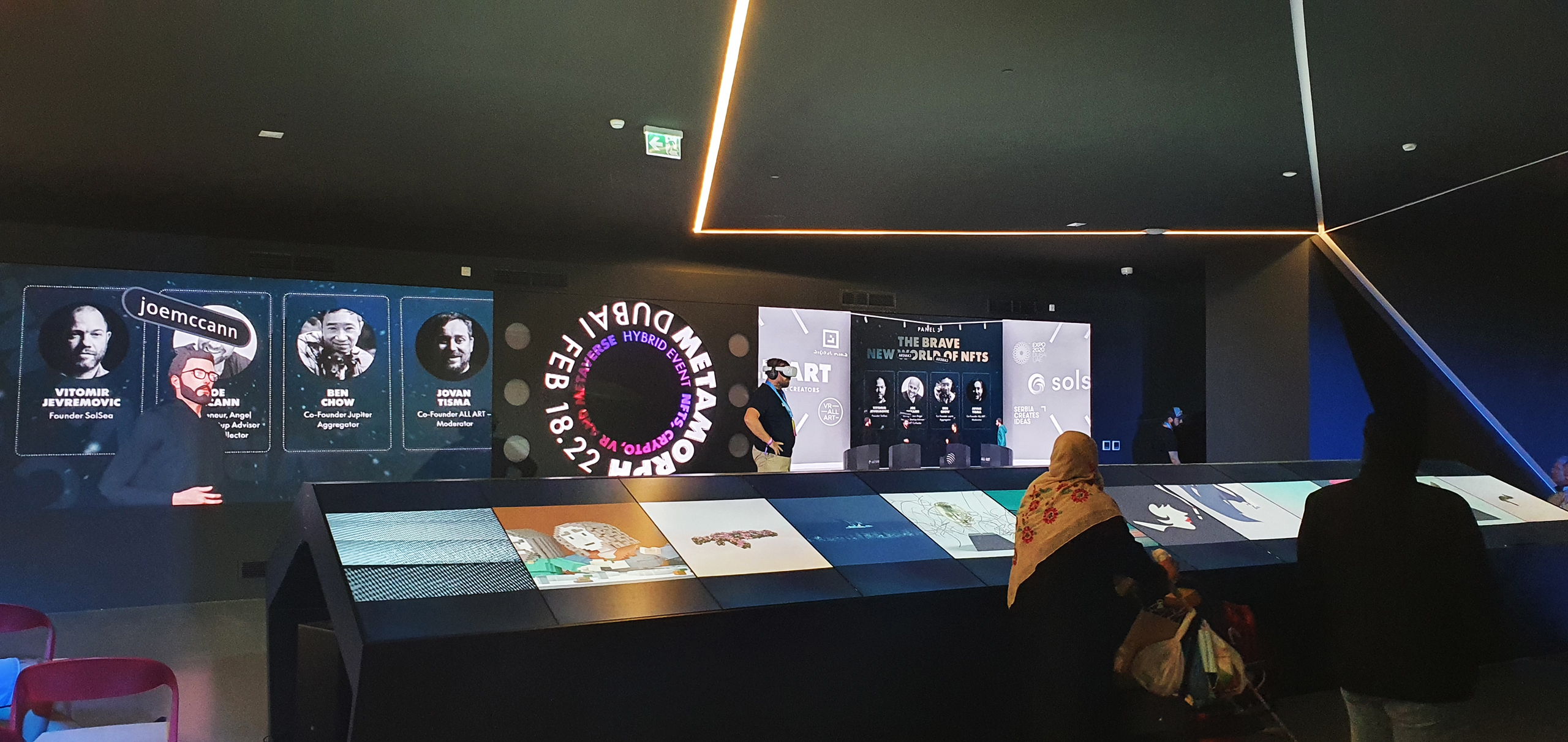 Inside the event, a person wearing a VR headset stands before a large screen surrounded by Metamorph branding. Below, an interactive display features a series of digital artworks, symbolizing the event's focus on NFTs and the Metaverse. This setting exemplifies Digital Mind's integration of live and virtual conferencing, enabling global real-time participation.