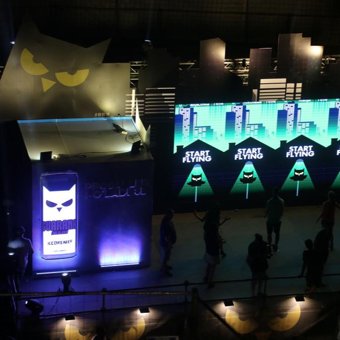 Overhead view of a darkened event space with a large LED wall displaying the Guarana logo and an interactive owl game, with participants and onlookers gathered around in a dimly lit atmosphere.