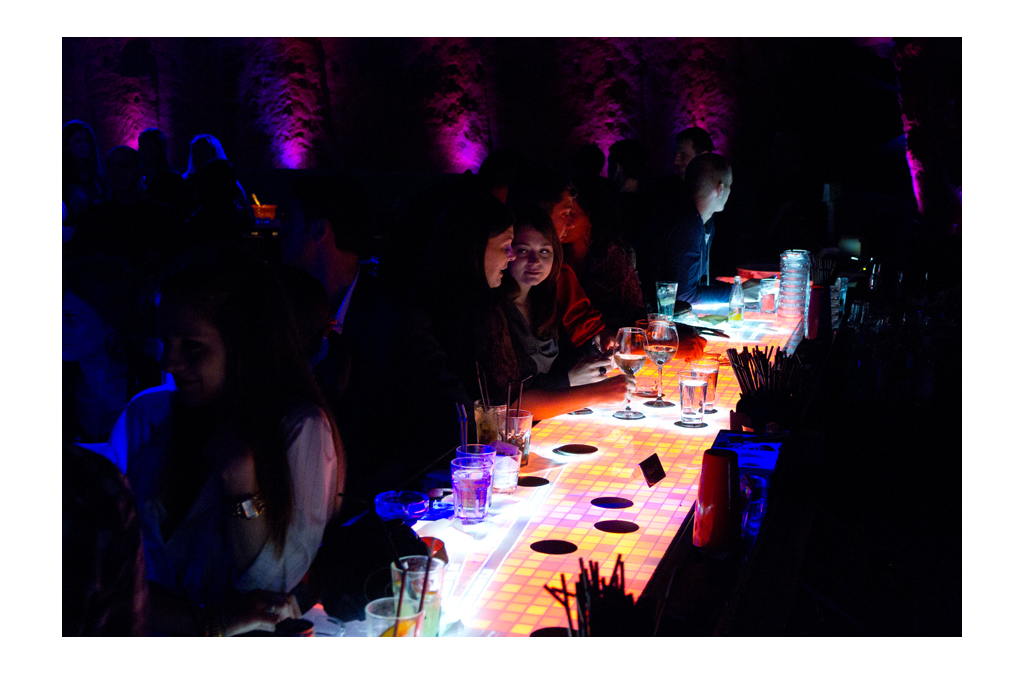 Patrons are seated at a vibrant, interactive bar counter with a continuous 6-meter surface that displays dynamic, touch-reactive visuals. The bar's unique technology allows for object detection and interaction, adding to the immersive experience.