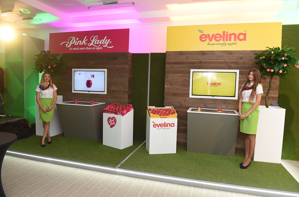 Two promotional booths for apple brands "Pink Lady" and "Evelina" with large brand logos, staffed by representatives in branded attire, and featuring display screens for interactive learning about the products.