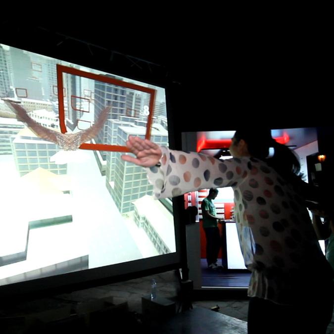 A person simulating eagle flight with arms spread in front of a large screen displaying a virtual 3D cityscape, engaging with an interactive flying game using a Microsoft Kinect sensor.