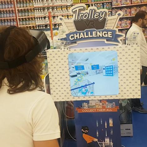 A participant is engrossed in the 'VR Trolley Challenge', wearing a virtual reality headset and standing in front of a promotional display in a supermarket. The screen above shows a 3D shopping environment where the game takes place, set against a backdrop of busy supermarket aisles.