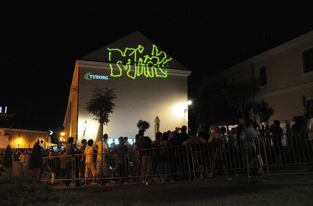 A nighttime view of a crowd gathered around a building, watching a laser painting in action. The wall is lit up with a neon green laser-painted word, offering a modern twist to public art through interactive technology.