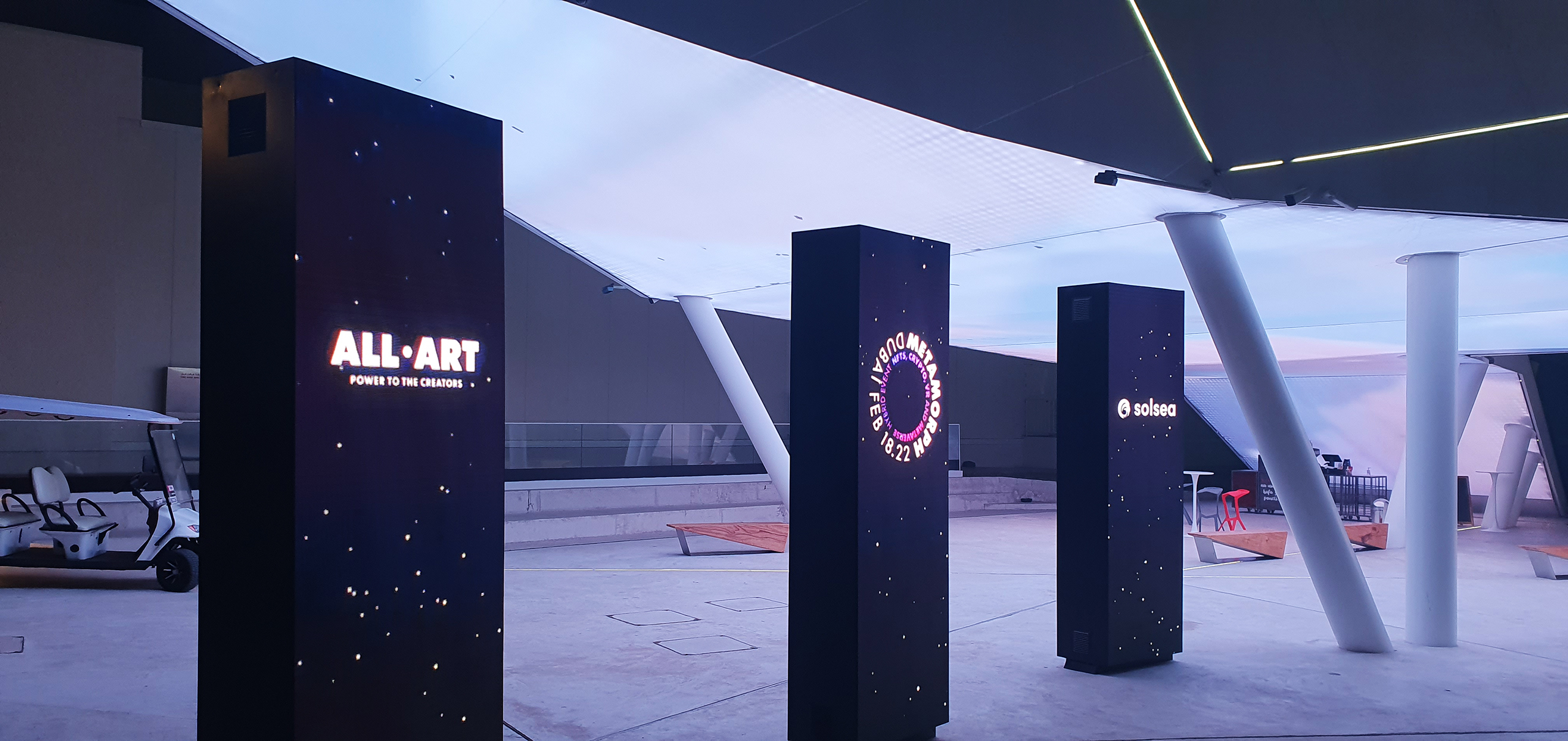 An installation features multiple illuminated pillars with branding for the ALL-ART and Solsea platforms, part of the Metamorph event by Digital Mind and VR-All-Art. This setup showcases the innovative avatar system intended to transform event experiences, highlighting the fusion of physical and virtual worlds in the context of crypto and NFTs.