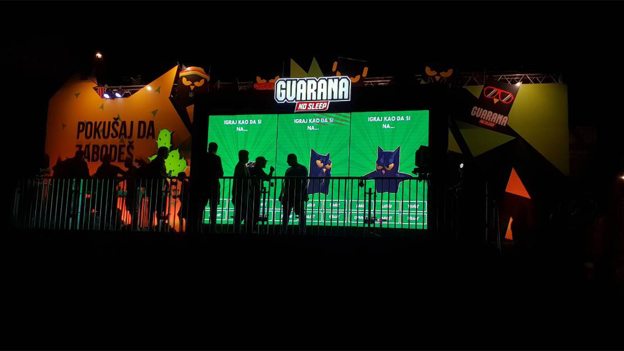 Festival-goers gather in front of a vibrant stage setup at the Guarana No Sleep area during EXIT Festival, engaging with an interactive game displayed on a giant screen, adding a digital interactive element to the music event.