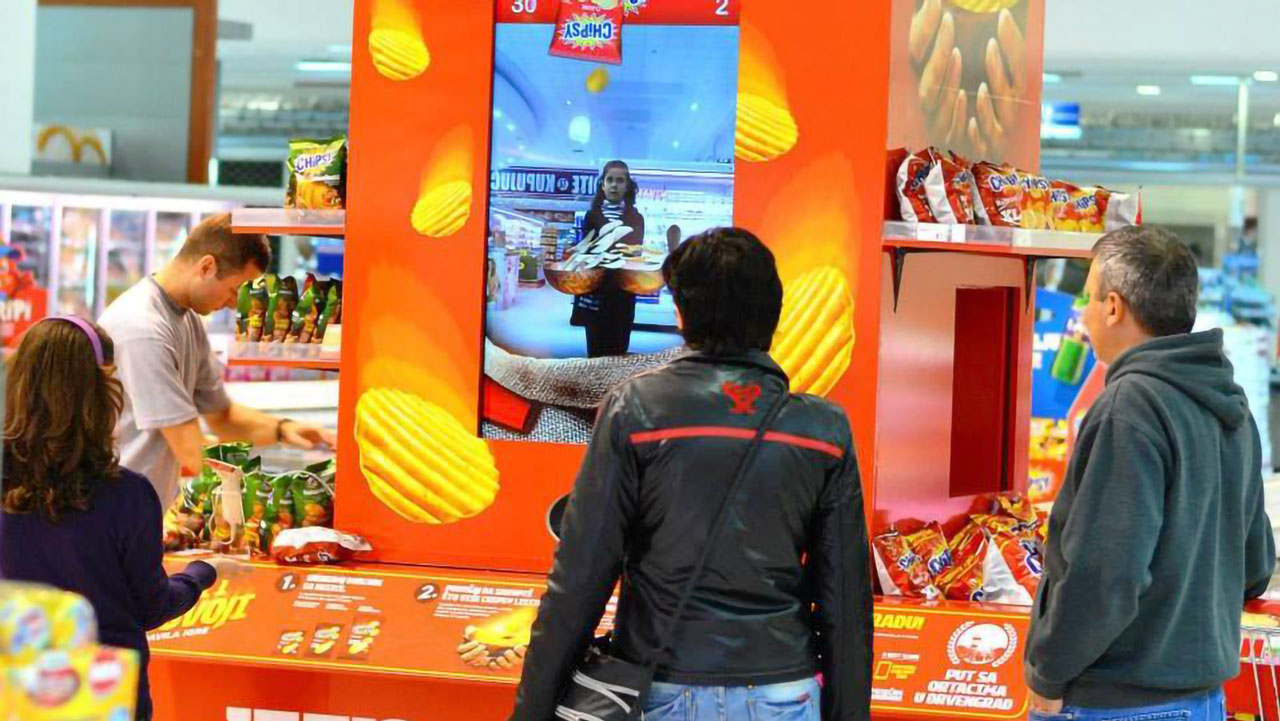 Shoppers watching a player interact with a large Chipsy brand augmented reality game installed in a grocery store.