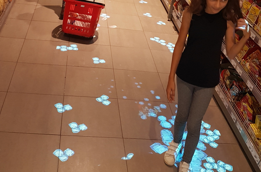 A child stands on a supermarket floor with blue flower projections that move and change with her steps.