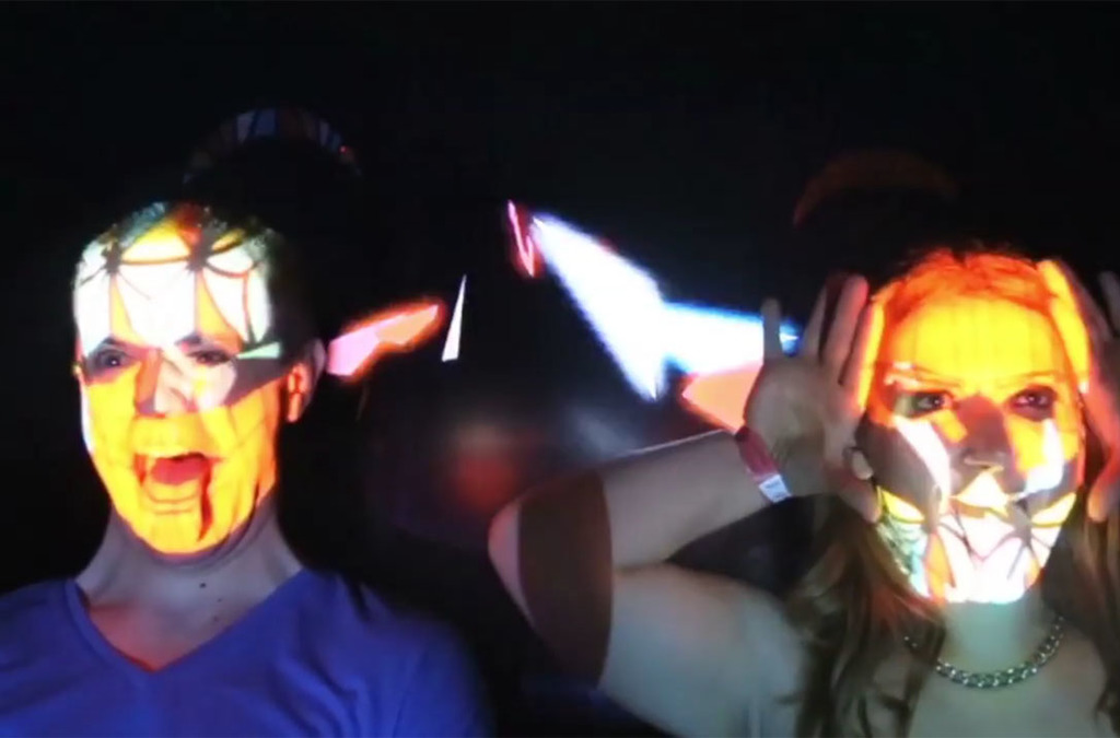 A real-time face mapping display showing two individuals with animated geometric patterns on their faces, resembling vibrant masks.