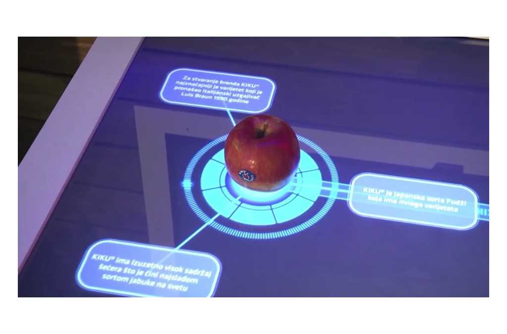 An interactive console screen displaying information about an apple placed on its surface. The screen shows circular graphics and text bubbles around the apple, providing details about the fruit’s variety and characteristics.