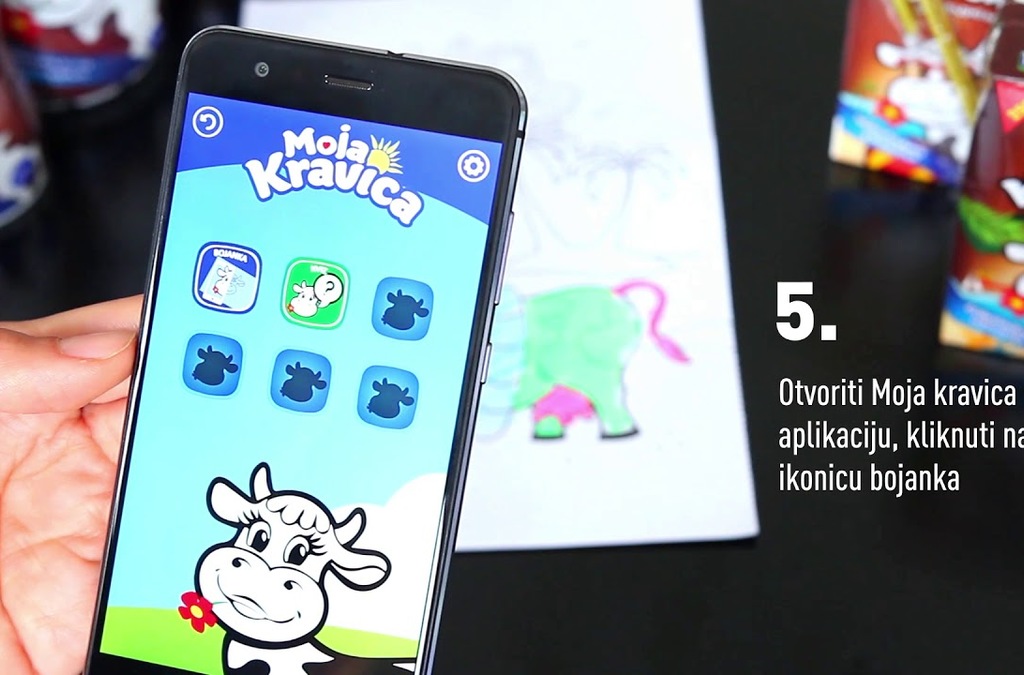 A smartphone screen displaying the 'Moja Kravica' app interface with icons for various activities, held over a child's drawing, showcasing the app's functionality in a playful setting.