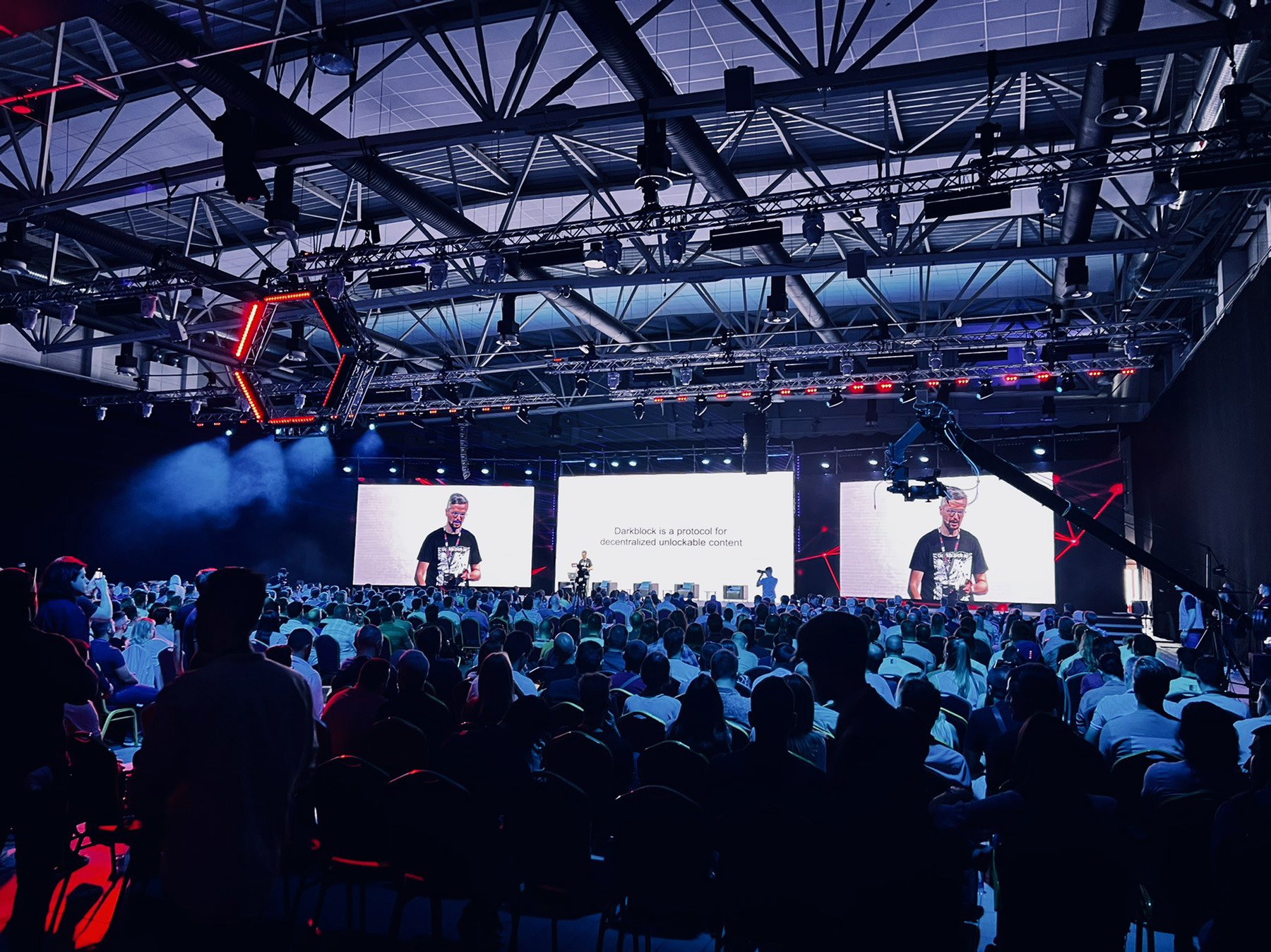 A speaker at the TMRW Conference presents on the topic of decentralized unlockable content, with a large attentive audience and a backdrop of the conference's branding and futuristic red and blue lighting.