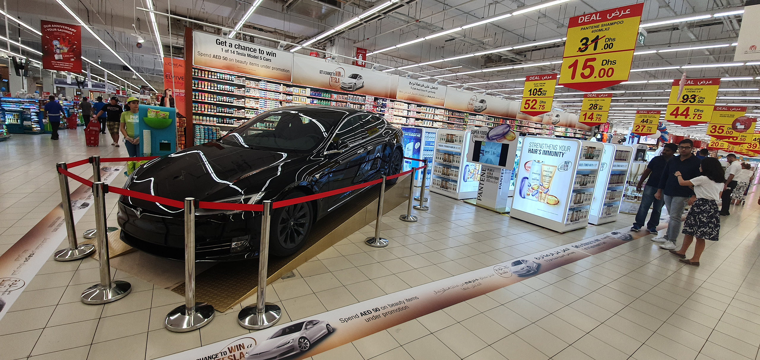  sleek black car displayed in a supermarket with promotional banners overhead. The car is cordoned off with red velvet ropes on metal stanchions, and a banner advertises a chance to win this model with a certain amount of spend on beauty items. Shoppers are seen in the background browsing aisles with various products on sale.