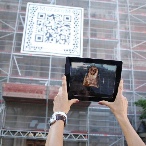 A person uses a tablet to scan a special ARQR code from the MUZZEUM project displayed on a building, revealing a virtual artifact on their screen.