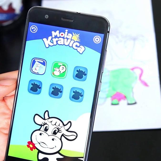 A smartphone screen displaying the 'Moja Kravica' app interface with icons for various activities, held over a child's drawing, showcasing the app's functionality in a playful setting.