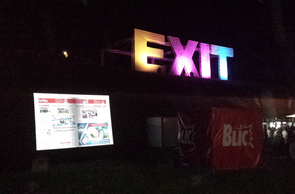 A nighttime scene of an outdoor digital installation where a large projected newspaper page is displayed next to the glowing "EXIT" sign, demonstrating the Gesture Page Flipping system.