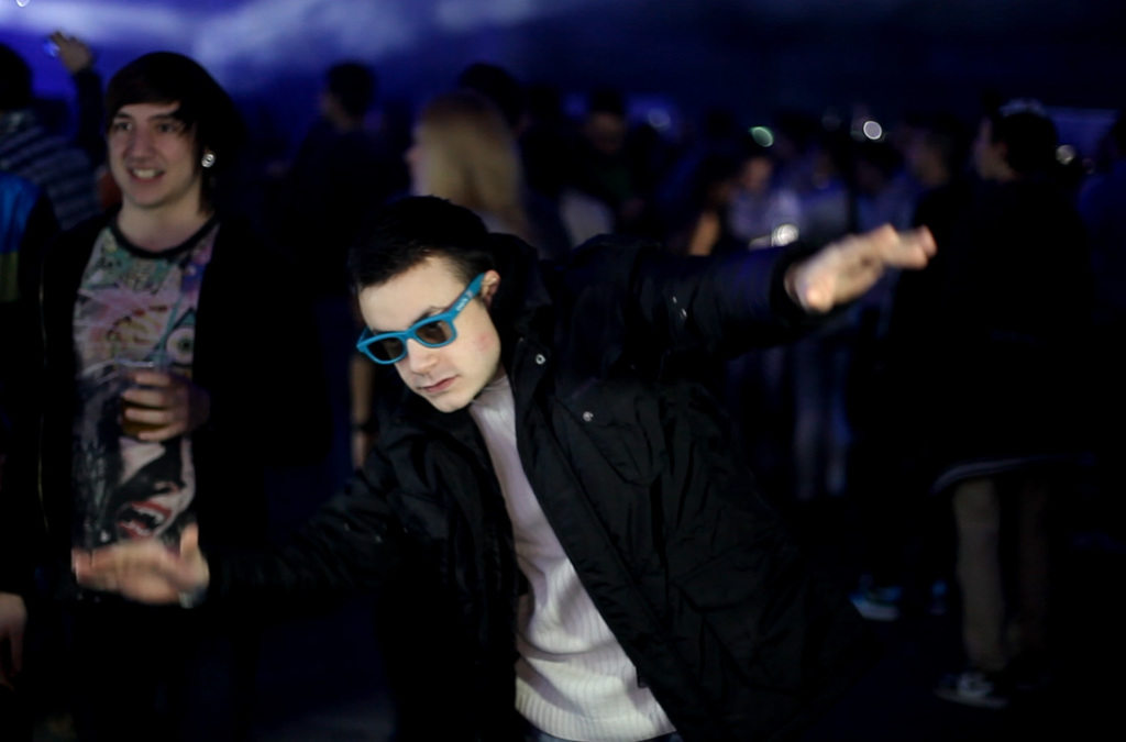 A person with outstretched arms leaning forward, imitating flight at a nighttime party with a crowd and colorful lights in the background.