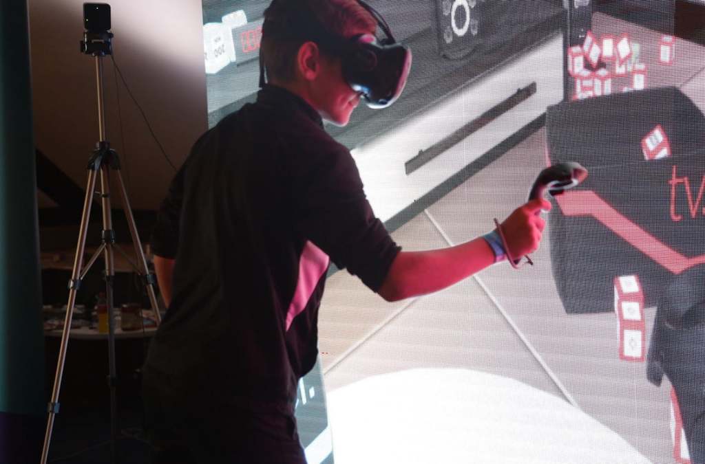 A person with a VR headset and handheld controller actively engages in a VR game at the Balkan TUBE Fest, with a tripod-mounted camera capturing the action and a game interface projected in the background.