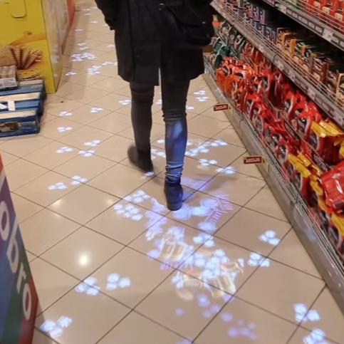 A person walks through a supermarket aisle with interactive blue flower projections on the floor, following their movements.