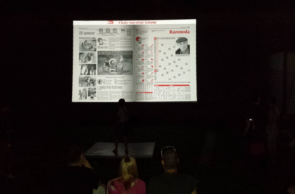 A silhouette of a person flipping through a projected digital newspaper page using hand movements detected by Microsoft Kinect technology in a low-lit setting.