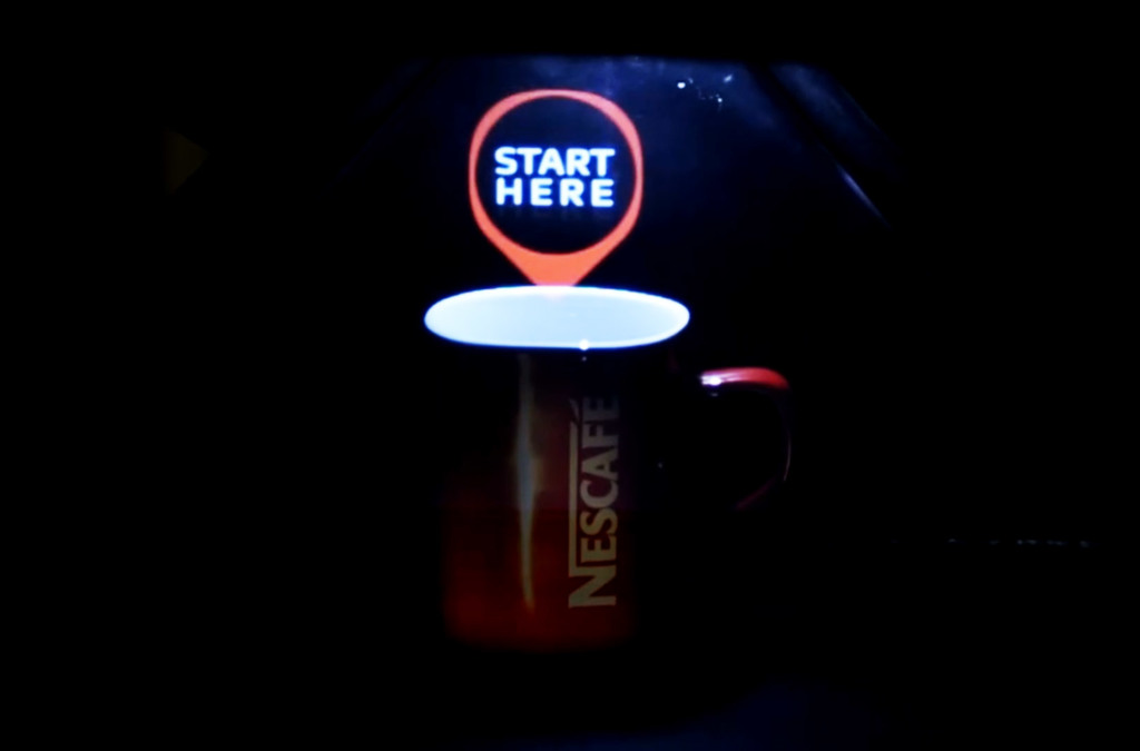 The same Nescafe cup within the hologram pyramid, now displaying an orange "START HERE" holographic sign above it, illustrating an interactive holographic presentation.