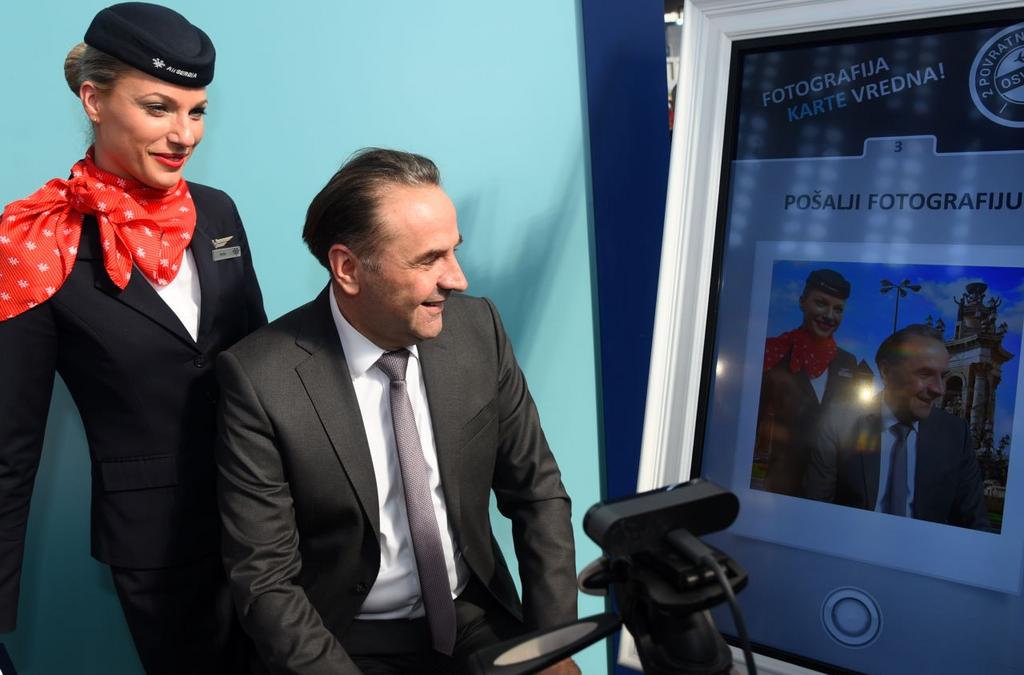 An Air Serbia representative stands next to a man interacting with a digital kiosk, sending a virtual postcard, part of the airline's promotional activities at the Tourism Fair.