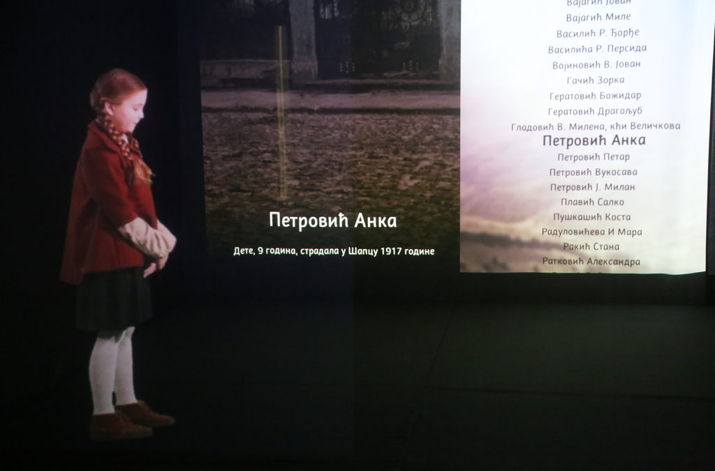 A young girl stands in front of a life-sized holographic projection displaying names and a historical scene, part of an interactive museum exhibition about WWI.
