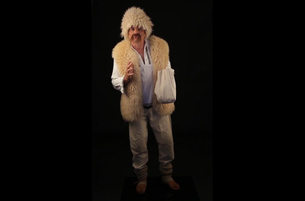 A man dressed in traditional attire with a white fur hat and vest, holding a sling bag.