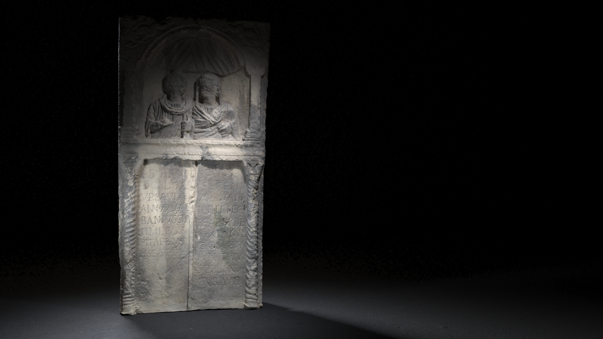 An ancient Roman funerary relief depicting two figures seated inside an arched niche, above a weathered Latin inscription, set in a tall vertical stone slab under dramatic lighting against a dark background.