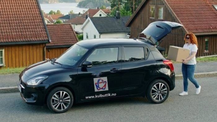 Et bud som bĂŠrer flere esker fra en hvit varebil med kulbuttik.no-logo foran et bolighus.