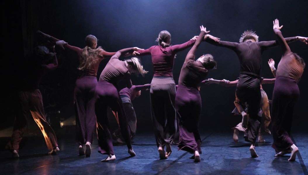 Group of dancers dressed in burgundy dancing in a circle on a dark stage, their backs are to the camera