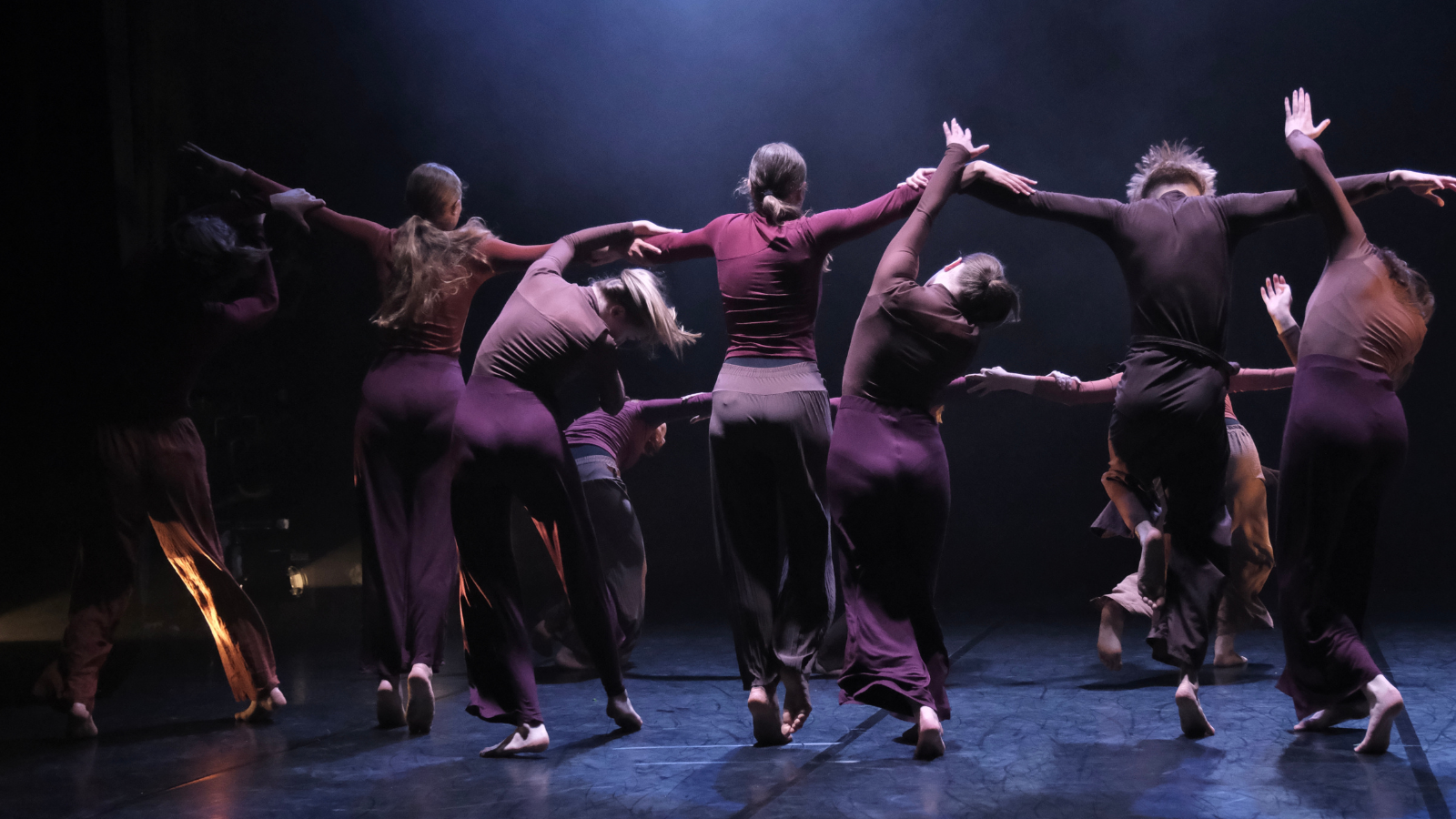 Group of dancers dressed in burgundy dancing in a circle on a dark stage, their backs are to the camera