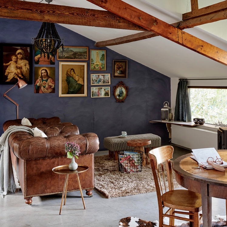 Woonkamer onder schuin dak met bruine Chesterfield bank, blauwe wand en schilderijen