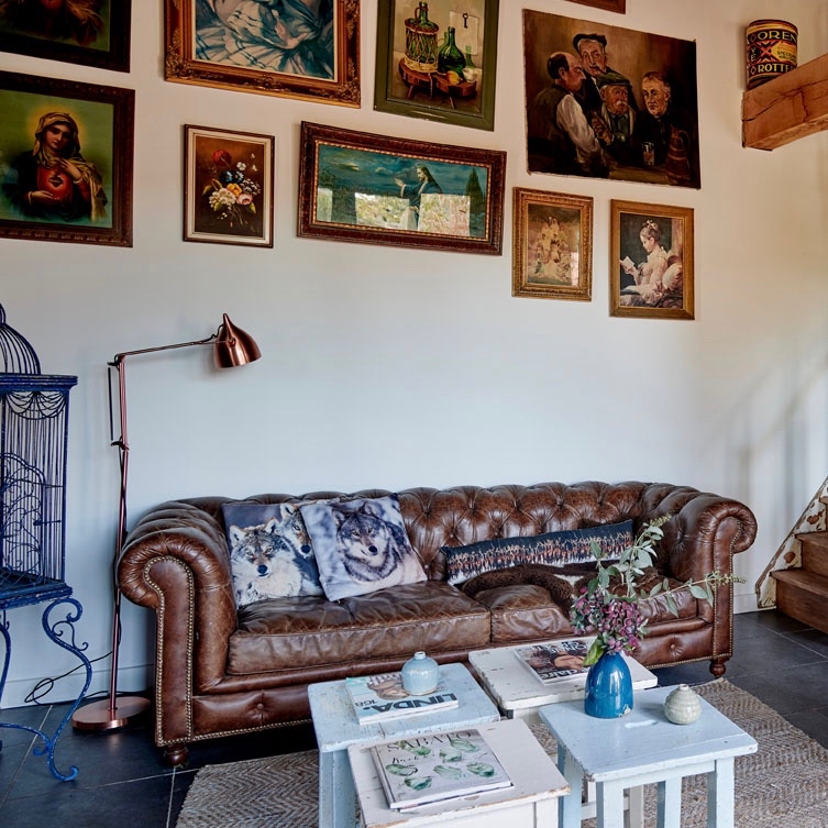 Zithoek met bruine leren Chesterfield en losse salontafels, diverse schilderijen erboven