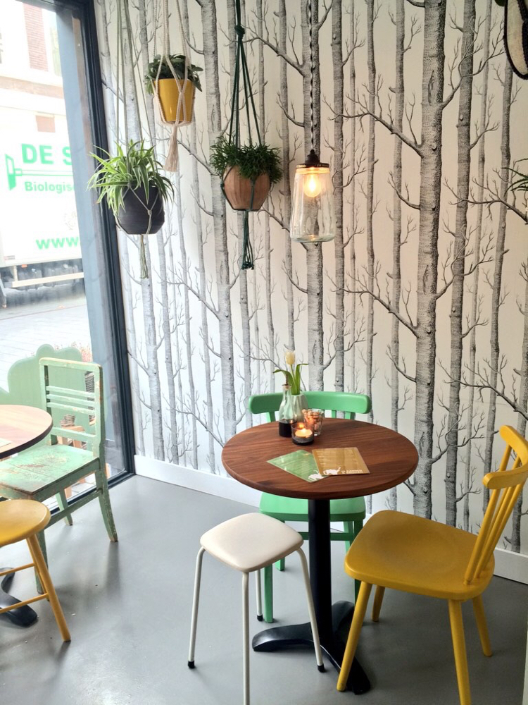 Ronde tafels voor raam aan straat met 2 stoeltjes en achterwand behang van bomen en hangende planten