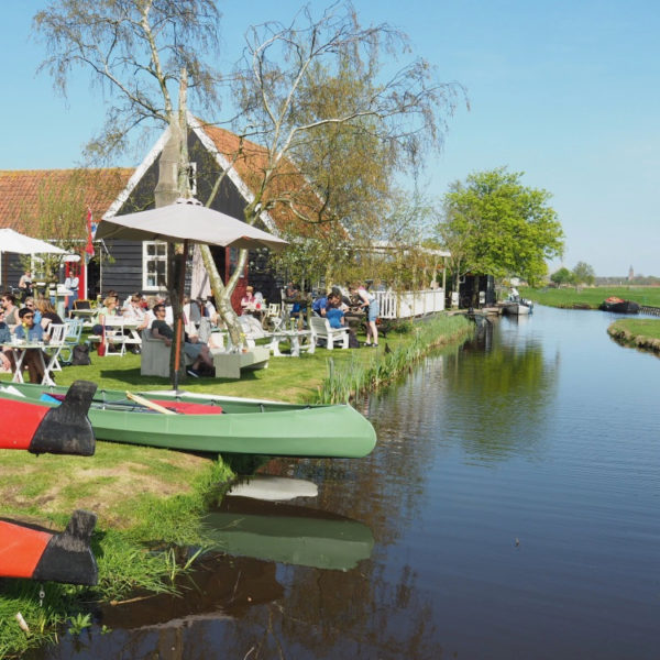 Een brede sloot met kano's in het gras en terrasstoelen met parasols