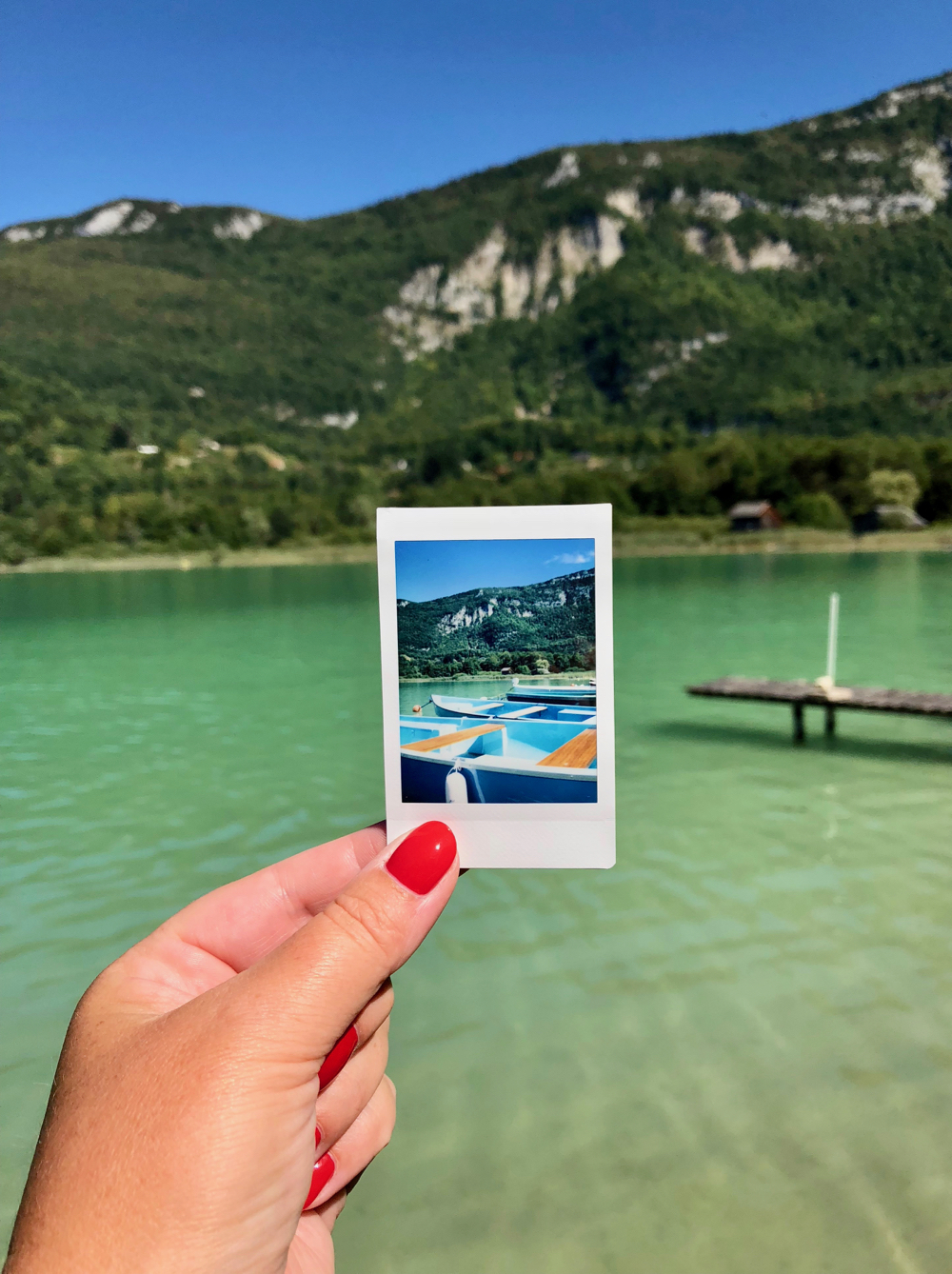 Hand met rode nagels houdt een polaroid vast voor een bergmeer met groen water