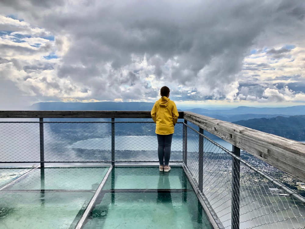 Een uitkijkpunt met glazen bodem met daarop een meisje met een gele regenjas die naar de bergen staart