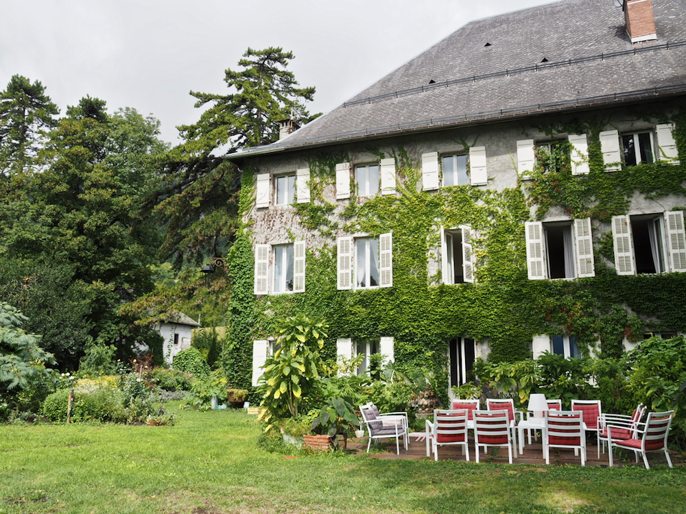 Buiten aanzicht van Chateau des Allues. Een gevel met grote ramen, veel groen eromheen in de tuin en een zittje