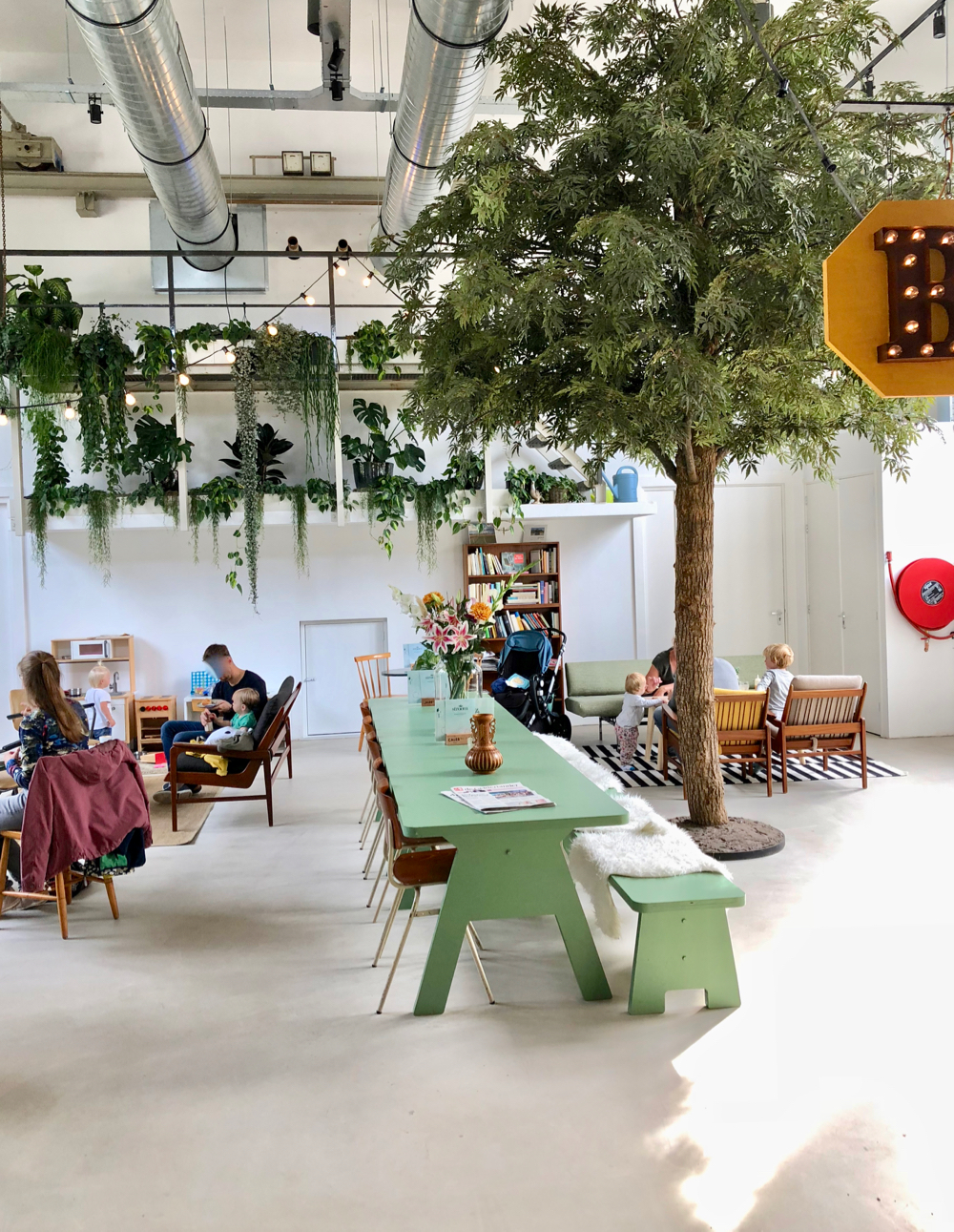 Een hoge ruimte bij Stroom Lent, met lange groene tafel met banken, een boom en veel planten aan de muur