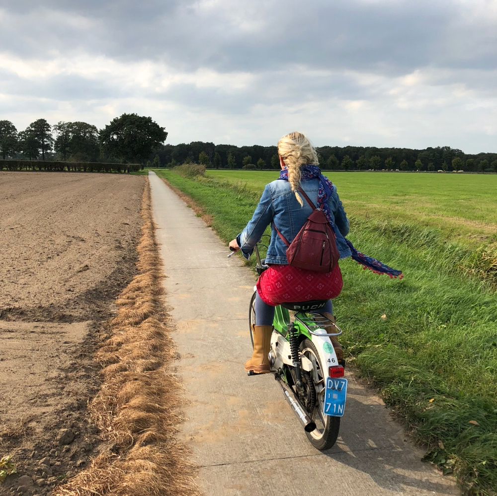 Toffe Puch met dame met lange blonde vlecht op een fietspaadje door de weilanden