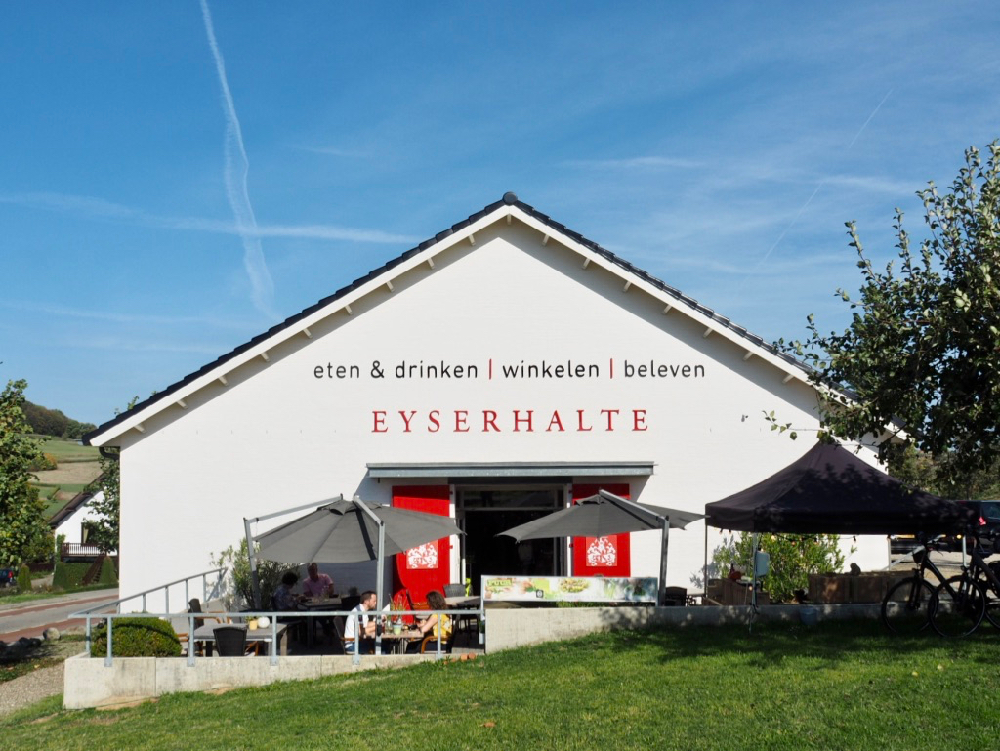 Vooraanzicht van lunchwinkel en delicatessenzaak de Eyserhalte. Een witte gevel met rode deuren en terras ervoor.